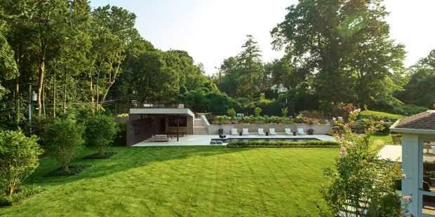 A landscaped residential backyard with a broad manicured lawn, mature trees, and a modern low-profile outdoor entertaining structure. The structure includes a covered seating area, broad pale paving, a raised terrace with steps, and multiple outdoor loungers. A small portion of another building is visible at the right edge. The setting is green, private, and garden-oriented, photographed in warm daylight.