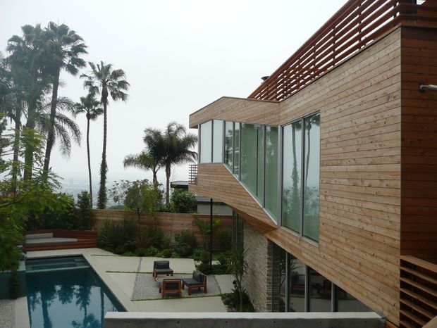 Exterior view of a contemporary multi-level residence clad in horizontal timber boards, with extensive floor-to-ceiling glazing and a rooftop terrace screened by horizontal timber slats. A landscaped courtyard includes a rectangular swimming pool, paved outdoor seating area, and tropical palms.
