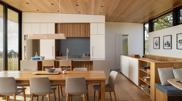 A bright, open-plan contemporary kitchen and dining area with extensive timber detailing. The room features a long timber dining table, upholstered dining chairs, a compact kitchen with dark tiled splashback and timber cabinetry, a timber-lined ceiling, large windows, and built-in timber bench seating with shelving along the right wall.