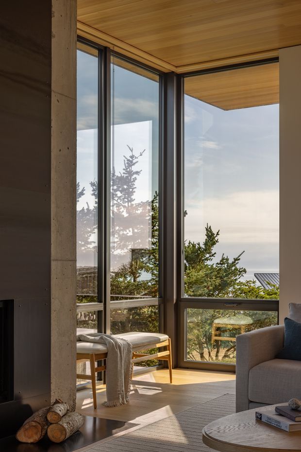 A warmly lit contemporary living room with full-height corner glazing overlooking trees and a distant coastal or open landscape. The room features a timber-lined ceiling, polished concrete structural column, dark built-in fireplace wall, timber flooring, a light upholstered armchair and sofa, and a round stone or concrete coffee table. Strong indoor-outdoor connection and abundant natural light define the space.