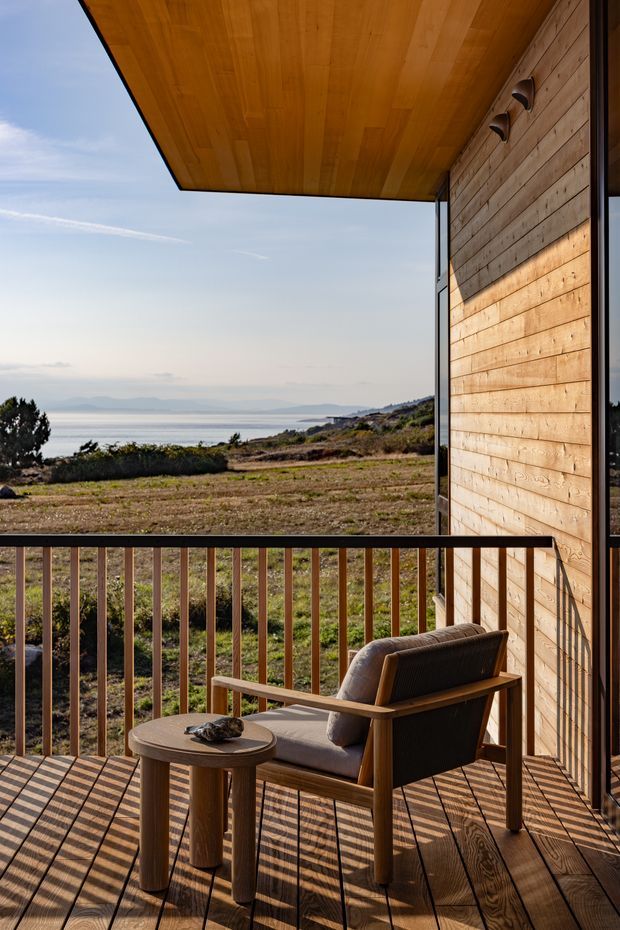A compact timber-clad balcony or terrace overlooking a coastal bay and rugged landscape. The space features a warm wood ceiling soffit, horizontal timber wall cladding, timber decking, slim dark metal framing and balustrade, and a simple wooden lounge chair with a small round side table. Strong sunlight creates pronounced linear shadows across the deck, with expansive views toward water and distant hills.