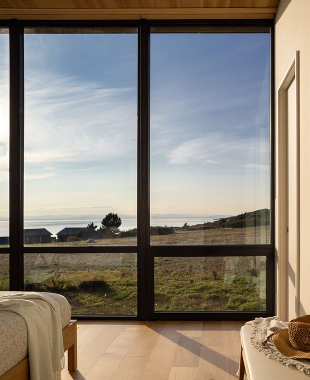 A serene bedroom corner with floor-to-ceiling dark-framed glazing overlooking a coastal landscape and ocean. The room has warm timber ceiling boards, pale wood flooring, cream walls, and a partially visible bed and upholstered bench, with strong natural light and expansive views.