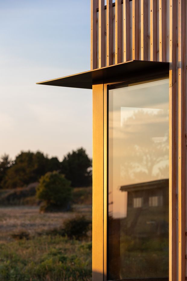 Close-up exterior architectural detail of a contemporary building at sunset, showing a large glazed opening with a dark metal projecting canopy and vertical timber sun-screen battens. The glazing reflects the surrounding landscape and another roofline, while the rural vegetation remains in the background.