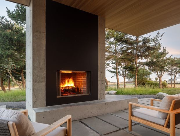 A partially enclosed outdoor lounge beneath a broad timber-lined roof canopy, centered on a built-in open fireplace set into a dark feature wall with exposed concrete columns and hearth bench. Timber-framed cushioned armchairs face the fire, while large openings frame a grassy, tree-lined landscape and distant buildings. The space has a warm, relaxed indoor-outdoor character with strong concrete, timber, and fire elements.