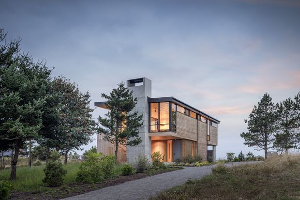 Exterior architectural photograph of a contemporary two-storey house set within a landscaped, coastal-looking site. The home has a compact rectilinear form, a tall concrete-like vertical tower with a rooftop opening, extensive timber-toned horizontal cladding, dark-framed glazing, and a covered upper-level balcony or recessed glazed corner. A paved path leads toward the entrance, with mature trees and low planting surrounding the building under an overcast evening sky.