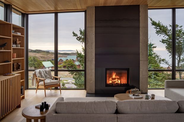A warm, contemporary living room with a large pale sectional sofa oriented toward a dramatic central fireplace. Floor-to-ceiling glazing provides expansive views of a hilly, wooded setting and nearby homes. The room features a timber-lined ceiling, built-in timber shelving and cabinetry, a dark stone or slate fireplace surround, pale flooring, and a sculptural mid-century-style lounge chair. The composition is calm and refined, with strong indoor-outdoor visual connection.