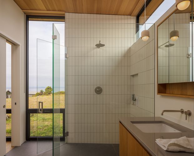 A contemporary bathroom with a walk-in glass shower, full-height cream grid tiles, dark grey floor tiles, timber ceiling, and warm timber vanity cabinetry. A large mirror reflects the vanity and pendant lights, while a glazed door opens toward a grassy coastal-looking landscape.