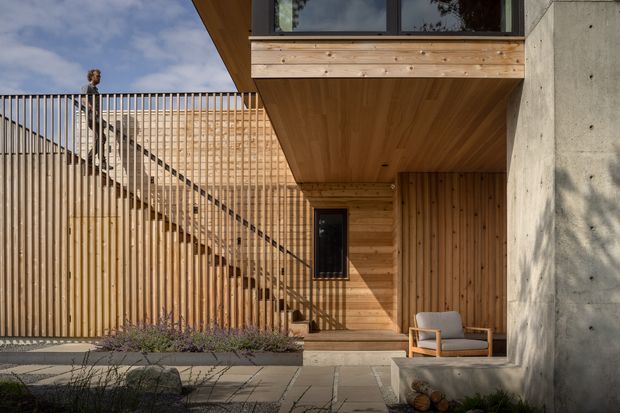 A modern residential courtyard or entry area with warm vertical timber screening, an exterior timber staircase and balcony soffit, exposed concrete walls, paved landscaping, and a small outdoor seating nook. The composition emphasizes the building facade and sheltered outdoor space, with strong geometric lines and natural materials.