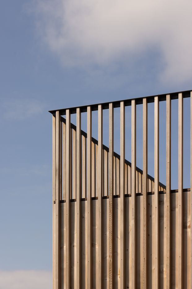 Close-up exterior architectural detail showing a rhythmic timber facade or screen composed of vertical wooden battens fixed to a darker horizontal roofline or beam. The battens vary in height along a sloping profile, creating a strong geometric silhouette against a pale blue sky. The image is tightly cropped, with no visible interior or broader site context.