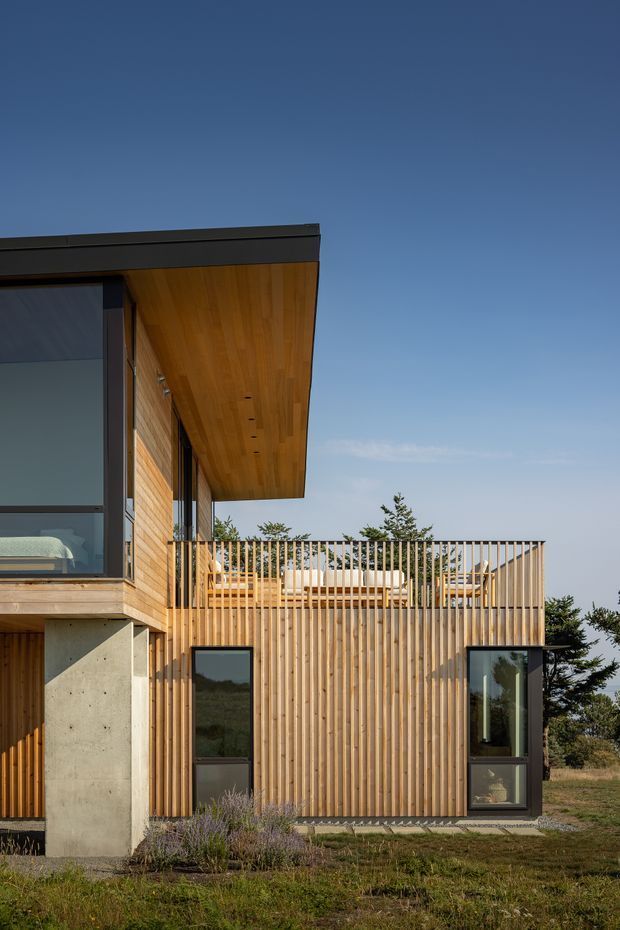 Exterior view of a contemporary timber-clad house with a projecting flat roof and warm timber soffit. The facade combines vertical timber screening and cladding with exposed concrete supports, large dark-framed glazing, and an elevated outdoor terrace with timber balustrades. Sparse coastal or rural planting surrounds the building under a clear blue sky.