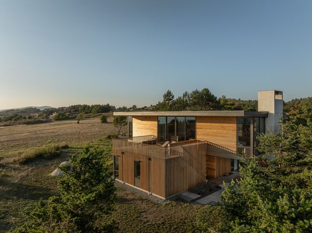 A modern two-storey timber-clad house set within a rural, lightly rolling landscape. The building has a low flat roof, extensive dark-framed glazing, a projecting upper-level terrace with timber balustrades, and a tall rectangular chimney or service volume. Dense shrubs surround the house, while open fields and distant hills extend behind it.