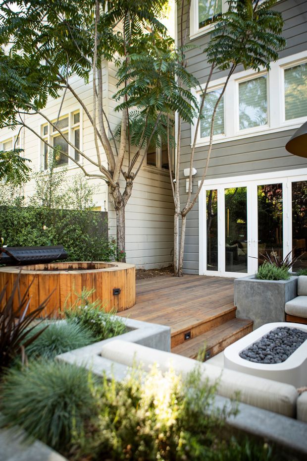 A landscaped outdoor courtyard or terrace adjoining a modern multi-storey home. The space features a timber deck with steps, a built-in round wood-clad spa or hot tub, concrete planters filled with ornamental grasses, outdoor seating, and a white firepit or coffee table filled with dark stones. The house has grey horizontal weatherboard cladding, white-framed windows, and glazed French doors opening onto the deck, with mature trees providing a leafy setting.