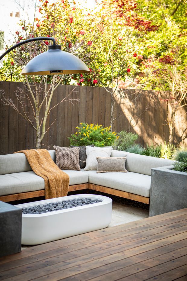 A landscaped outdoor lounge on a timber deck, featuring a curved pale upholstered sectional, low white pebble-filled fire pit or table, oversized dome pendant heater/light, concrete planters, cushions, and garden planting enclosed by a timber fence.