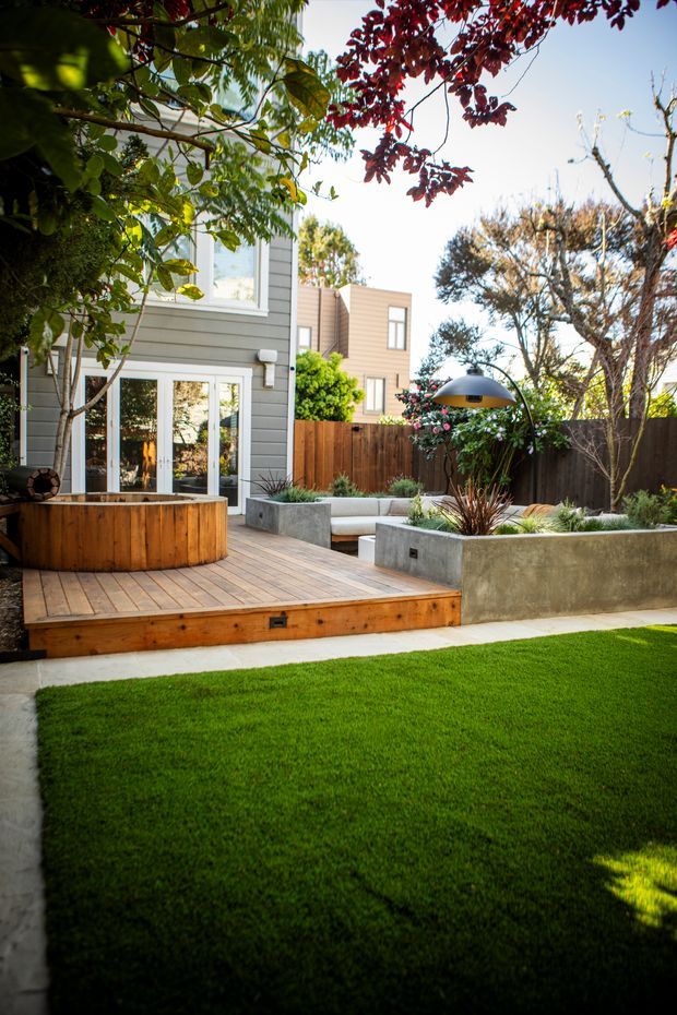 A landscaped residential backyard with a timber deck, built-in circular timber planter, concrete raised garden beds, built-in bench seating, a sculptural outdoor pendant light, timber fencing, and a neatly maintained lawn. The house has grey horizontal cladding and large glazed doors opening onto the deck, creating a contemporary outdoor entertaining space.