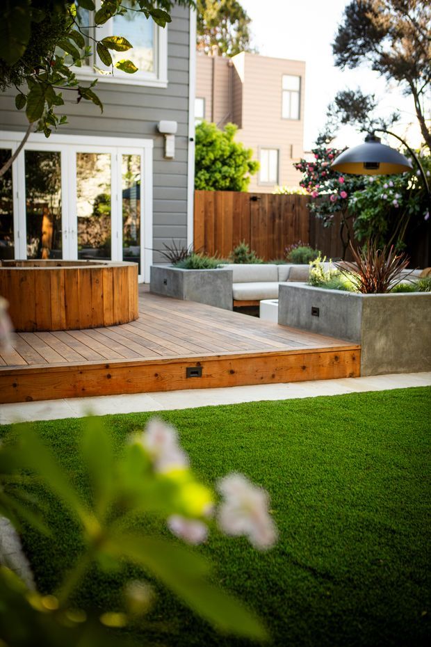 A landscaped residential courtyard viewed from the lawn, featuring a raised timber deck, built-in concrete planters and bench seating, a timber privacy fence, outdoor planting, and a large suspended outdoor light. A grey weatherboard house and neighbouring residential buildings form the backdrop.