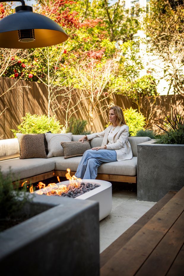 A landscaped outdoor lounge courtyard with a built-in upholstered sectional surrounding a contemporary gas fire pit. Concrete planters, timber decking, a timber privacy fence, and mature greenery create a warm, private entertaining area beneath a large suspended outdoor light.