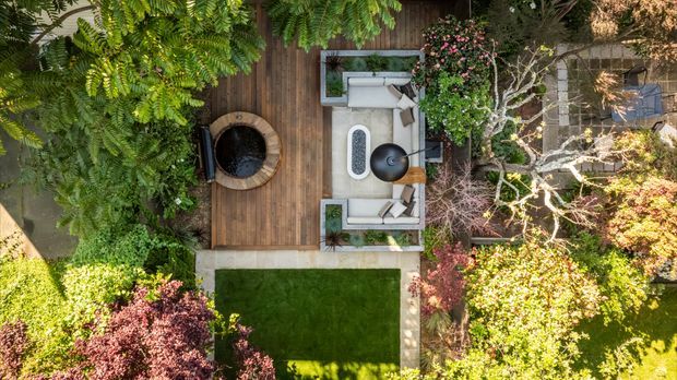 Aerial view of a landscaped residential outdoor entertaining area, with a timber deck, pale outdoor seating, a central fire pit or circular feature, and a neatly edged lawn surrounded by mature ornamental planting and trees.