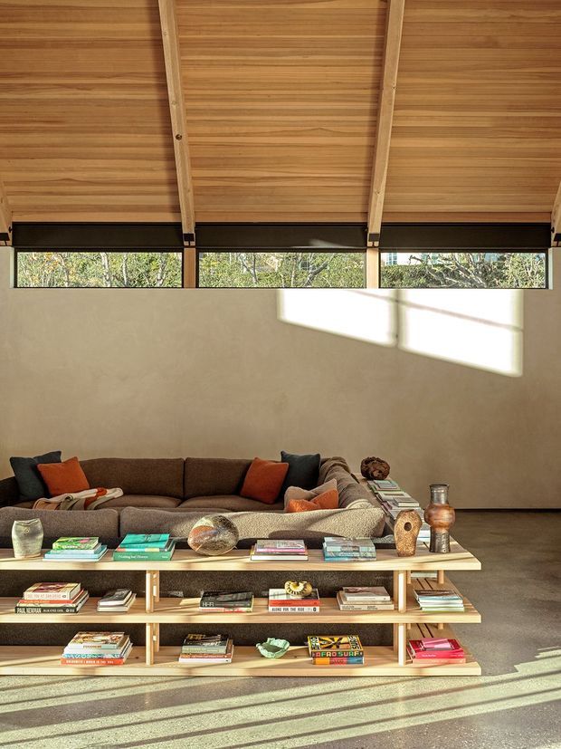 A warm, sunlit living room with a low brown sectional sofa, colourful cushions, and a long open timber shelving console filled with books and decorative objects. The room features a high sloping timber-lined ceiling with exposed pale timber beams, clerestory-style horizontal glazing, plastered walls, and a polished concrete floor. Strong sunlight creates geometric shadows across the wall and floor.