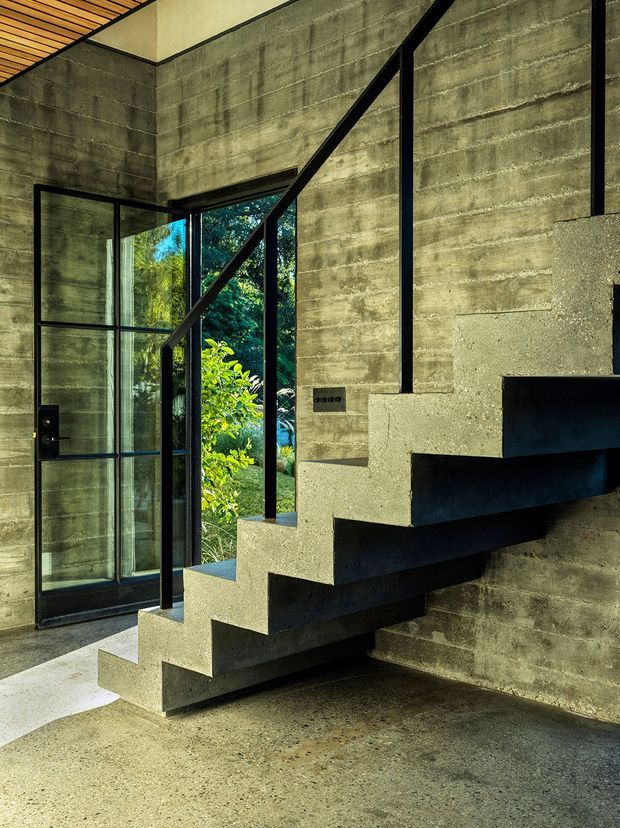 Interior view of a dramatic floating concrete staircase with black metal railing and exposed concrete walls. A black-framed glazed entry door opens toward lush greenery, while a warm timber slatted ceiling and polished concrete floor add contrast. The stair design is sculptural and minimalist with strong geometric lines.