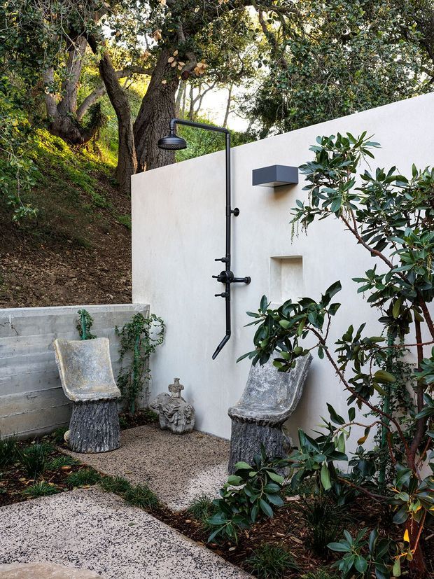 A secluded outdoor shower courtyard set against a white rendered wall and dense established planting. The black metal wall-mounted shower includes an overhead showerhead and handheld fixture, with a recessed wall niche and rectangular wall light. Two sculptural rustic stone or concrete chairs sit on a gravel surface beside textured concrete steps and a pale aggregate path. The setting feels natural, private, and resort-like.