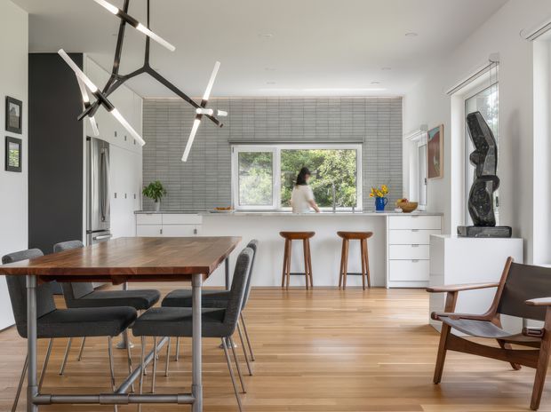 A bright open-plan dining and kitchen interior with a mid-century-influenced aesthetic. The room features a large timber dining table with upholstered dining chairs, pale timber flooring, white cabinetry, a grey horizontally tiled backsplash, stainless-steel appliances, and a white island or breakfast counter with two timber stools. A sculptural black linear pendant light hangs over the dining area, while a mid-century lounge chair and dark sideboard occupy the foreground and right side. Large windows bring in natural light and overlook greenery.