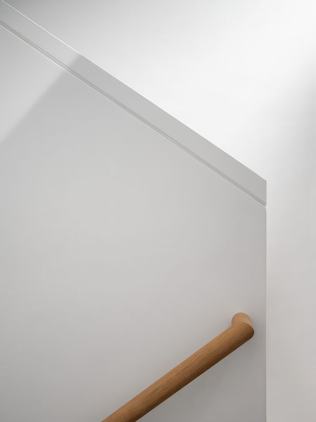 Close-up minimalist architectural detail showing a smooth white wall or door panel with a slim linear trim and a warm timber cylindrical handrail running diagonally across the lower portion. The composition is stark, clean, and highly abstract, with soft natural shadows and no identifiable room context.