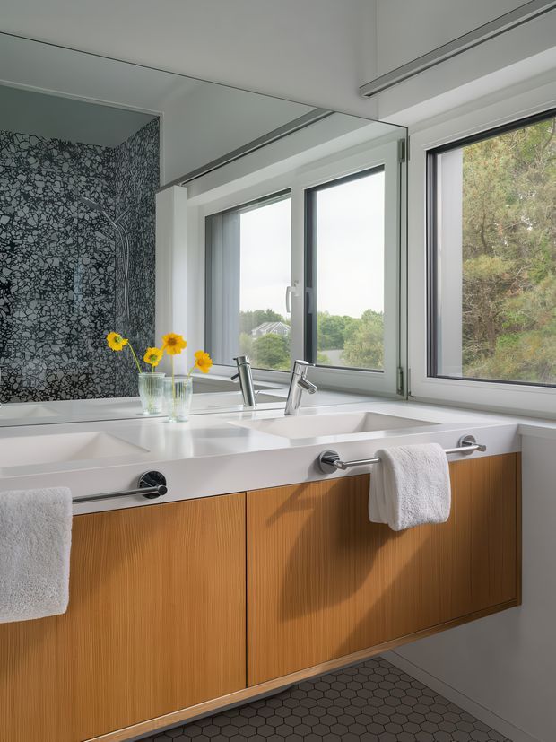 Bright contemporary bathroom vanity with a long white double basin benchtop, wall-mounted chrome faucets and towel rails, and warm vertical-grain timber cabinetry. Large windows provide abundant natural light and overlook trees, while a full-height dark patterned feature wall and dark hexagonal floor tiles add contrast. A large mirror reflects the vanity and windows.