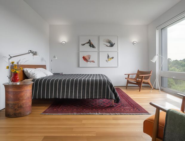 Bright, calmly styled bedroom with a low platform bed dressed in a dark striped blanket, a vintage wooden bedside drum table, framed bird artwork, a timber armchair, and a large patterned rug. The room has white walls, pale timber flooring, simple wall lights, and expansive corner glazing overlooking greenery.