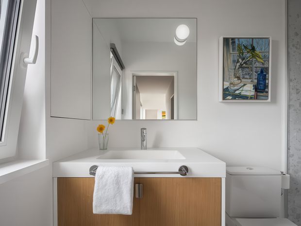 Bright, compact contemporary bathroom with a wall-mounted timber vanity, white integrated basin and chrome tapware. A large frameless mirror reflects the hallway, while a toilet, white-framed window, framed artwork and subtle patterned wall finish complete the space.