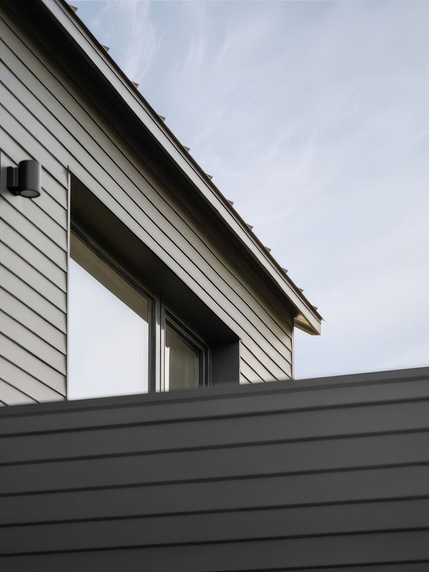 Close-up exterior architectural view of a contemporary house facade, featuring light horizontal weatherboard cladding, dark window and door frames, a deep roof eave, and a dark horizontal-clad foreground wall or fence. The composition emphasizes clean linear detailing against a pale sky.