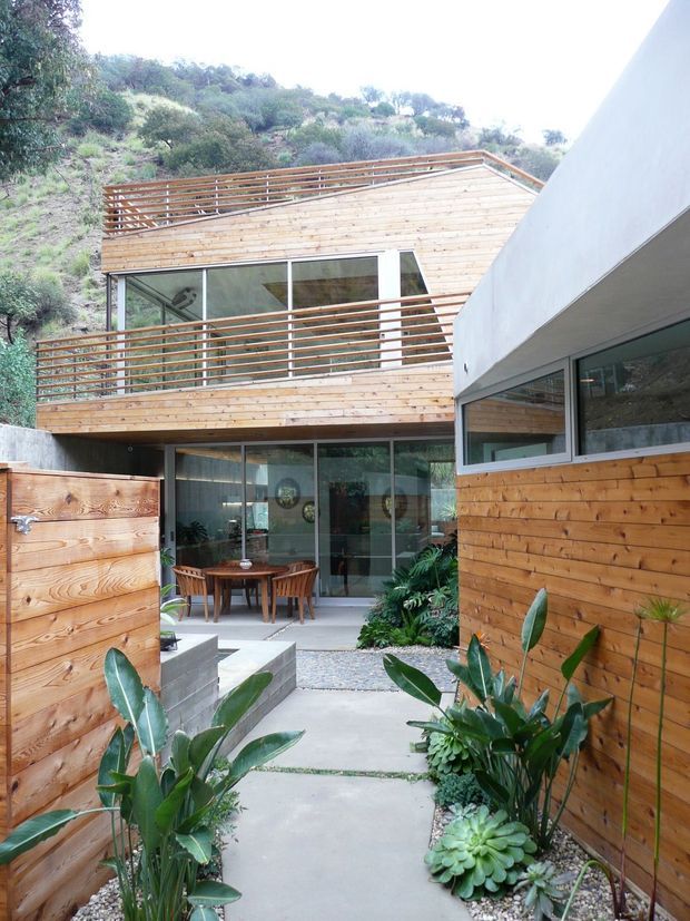 A modern multi-level residence built into a steep, landscaped hillside. The courtyard view shows extensive warm timber cladding and horizontal timber balcony railings, large aluminium-framed glazed doors and windows, concrete paving, and a dining area opening onto the outdoor space. Native and succulent planting softens the compact courtyard, while a second white-rendered building volume frames the right side.