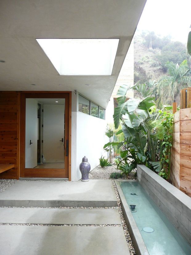 A modern covered entry courtyard with broad concrete stepping slabs, a timber-clad wall and glazed timber entry door, a large rectangular skylight, and a narrow linear water feature bordered by concrete blockwork. Dense tropical planting and a timber privacy fence create a lush enclosed setting.