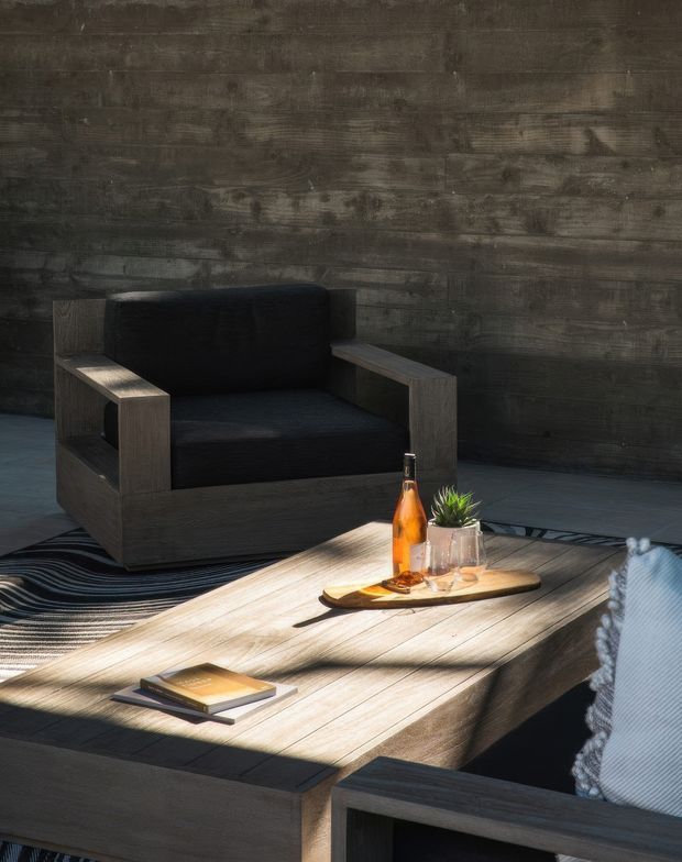 A moody, contemporary living room vignette featuring a dark timber-clad wall, a substantial dark wood armchair with black upholstery, and a large rustic timber coffee table. A patterned dark rug, small tray with bottle and plant, books, and a partially visible upholstered chair complete the relaxed lounge setting. Strong directional sunlight creates high contrast across the room.