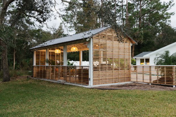 Exterior view of a compact single-storey pavilion or extension set among trees. The structure has a pitched metal roof, white posts and trim, extensive horizontal timber screening across the gable end and verandah, a timber deck, and warm pendant lights visible within the open-sided area. A larger white building is partially visible behind it.