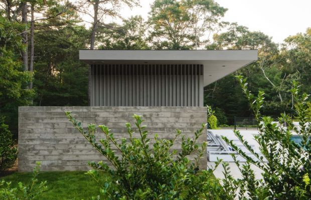 A modern pavilion-like structure set within dense greenery. The building has a long flat roof with a pronounced cantilever, a pale horizontal concrete-block wall, and a recessed upper section screened by closely spaced vertical slats. A light paved terrace or roof surface is visible to the right, partially obscured by shrubs.