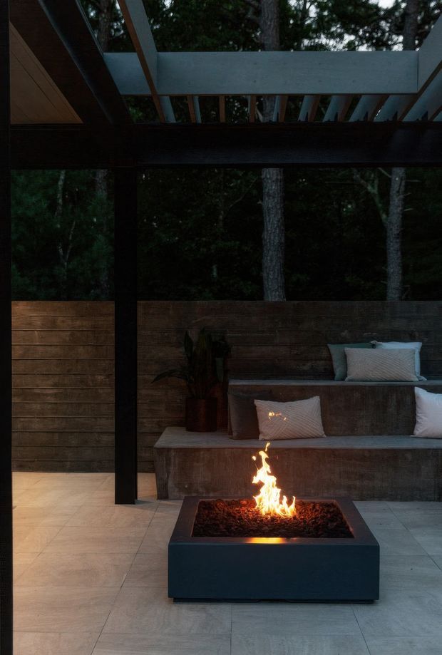 A moody covered outdoor entertaining area set among trees, featuring a contemporary square gas fire pit in the foreground, built-in timber-and-concrete bench seating with cushions, horizontal timber privacy screens, large-format grey floor tiles, and a partial pergola roof with exposed beams. The fire provides a warm focal glow against the dark forested setting.
