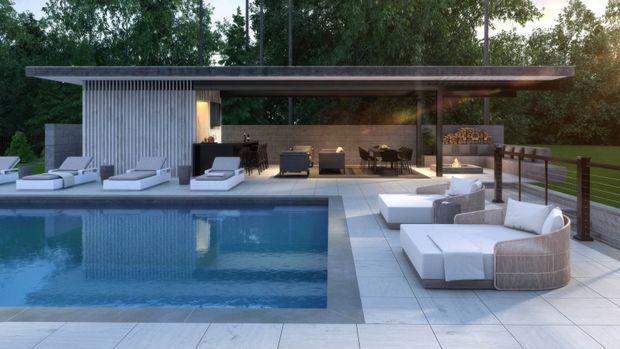 A refined modern poolside pavilion with a broad flat roof, floor-to-ceiling glazing, and a concrete or stone-clad vertical wall. The pavilion opens onto a tiled terrace furnished with white sun loungers and rounded outdoor seating, while a dark blue swimming pool occupies the foreground. Lush trees form a private backdrop, and the interior lounge and dining area are visible through the glazing.
