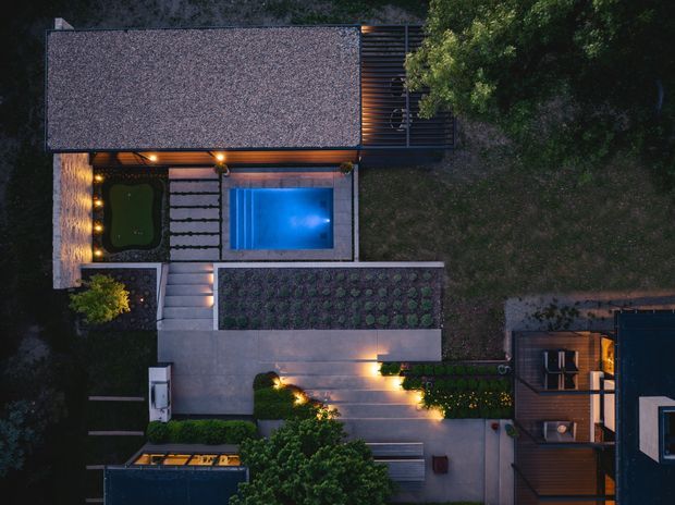 Aerial dusk view of a landscaped residential property showing a rectangular blue swimming pool, illuminated concrete pathways and terraces, planted garden beds, a small lawn area, and multiple connected roofed structures. The composition emphasizes the outdoor site layout, geometric hardscaping, and warm landscape lighting.