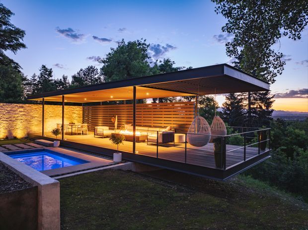 A modern elevated outdoor pavilion or poolside entertaining area at dusk, featuring a flat-roofed covered deck with slim dark structural posts, extensive glazing, warm timber slatted walls and ceiling, integrated seating, hanging egg chairs, and a small illuminated swimming pool. The structure overlooks a wooded landscape and distant horizon.