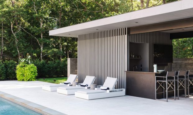 Contemporary poolside outdoor entertaining area with a flat-roofed pavilion, vertical slatted screening, integrated bar, bar stools, and white sun loungers arranged beside a swimming pool. The pavilion is set within a lush landscaped garden and uses a restrained palette of white, grey, charcoal, and natural greenery.