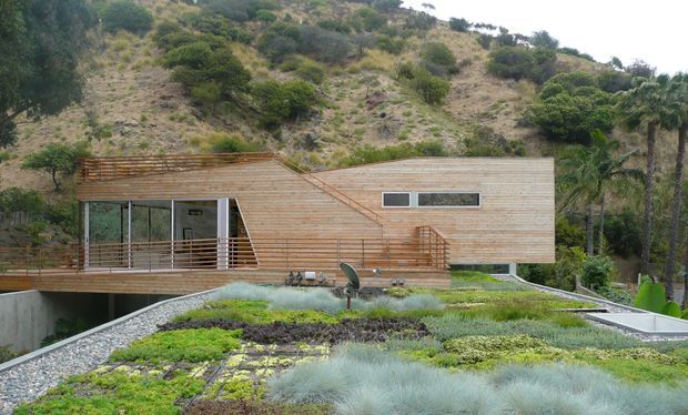 A contemporary hillside residence clad in horizontal timber weatherboards, with angular roof planes, extensive corner glazing, and a raised timber deck. The foreground features a planted green roof with gravel margins, while the building is integrated into a steep, densely landscaped site.