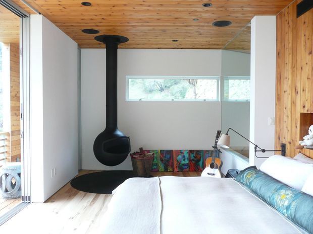 Bright contemporary bedroom with a low-profile bed, white bedding, pale timber flooring, and extensive timber wall and ceiling lining. A sculptural black suspended wood-burning fireplace is positioned centrally against a light wall, with high windows, sliding glazing, a guitar, and colourful artwork adding character. The room has a warm, airy timber-lined interior with strong indoor-outdoor connection.