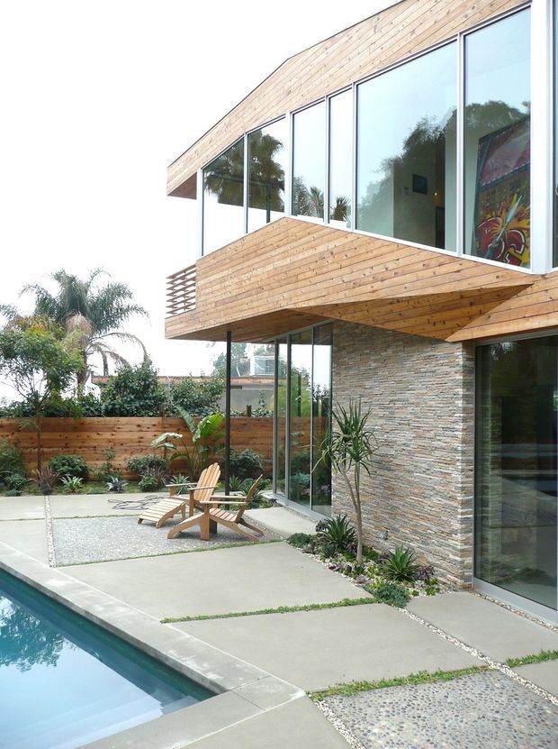 Exterior view of a contemporary multi-level residence surrounding a landscaped pool terrace. The building features extensive floor-to-ceiling glazing, projecting timber-clad volumes and balconies, a stacked-stone wall, concrete paving, and a pebble-textured walkway. Tropical planting, timber fencing, and outdoor lounge chairs frame the pool area.