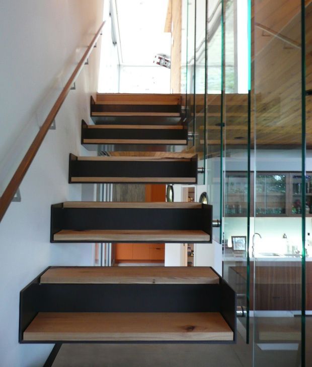 Interior view looking up a modern open-riser staircase with thick timber treads and dark painted stringers. A slim timber handrail runs along the white wall, while full-height clear glass partitions reveal a kitchen or utility area beside the stair. The composition emphasizes the floating stair construction, natural timber, black detailing, and abundant daylight.