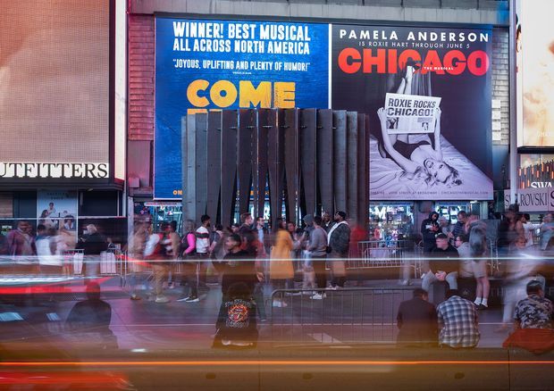 A lively urban commercial streetscape at night, with illuminated theatre and retail advertising, crowds of pedestrians, storefronts, and motion-blurred traffic in the foreground. The image has a vivid, energetic city atmosphere dominated by blue, red, orange, and dark tones.
