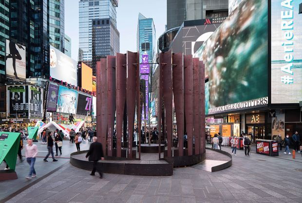 Urban public plaza surrounded by tall commercial buildings and dense illuminated advertising screens. The foreground features a large circular sculptural installation with vertical rust-coloured metal panels, surrounded by pedestrians on broad paved walkways. The scene has a contemporary metropolitan streetscape and high-rise commercial character.