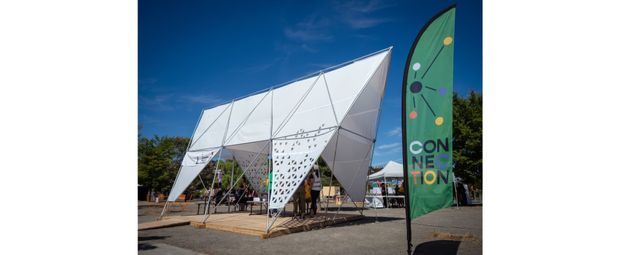 A temporary outdoor event pavilion with a striking faceted white tensile or panelled canopy, angular geometric supports, and perforated white screens. People are gathered beneath the structure, with a tall green convention banner in the foreground. The image is an exterior architectural/event-space view under a clear blue sky.