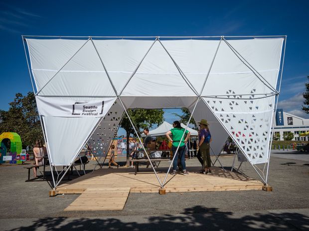 A temporary outdoor pavilion or event installation with a geometric triangular frame, white tensile fabric panels, and perforated white side screens. It sits on a timber platform in an open paved area, with people gathered underneath and a visible Seattle Design Festival sign. The structure has a lightweight, angular, modular appearance under clear daylight.