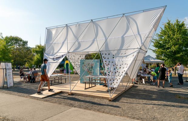 An outdoor temporary architectural pavilion with a dramatic angular steel frame and taut white fabric panels, creating a faceted canopy and perforated side screens. The structure sits on a raised timber platform in a public event setting, surrounded by picnic tables, people, trees, and adjacent temporary stalls under bright daylight.