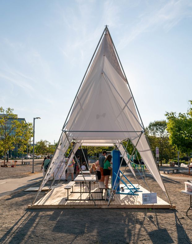 An outdoor temporary pavilion or event structure with a tall triangular, tent-like form. Translucent fabric panels are tensioned over a slender metal frame, creating a dramatic pointed canopy above a raised timber platform. People and event furnishings occupy the open interior, with paved public space, trees, and modern buildings in the background.